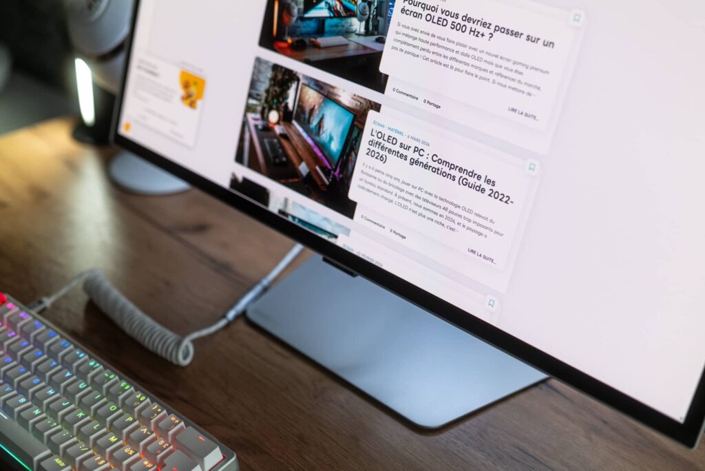 angled computer monitor showing tech articles on a desk with a rainbow-backlit keyboard and coiled cable nearby