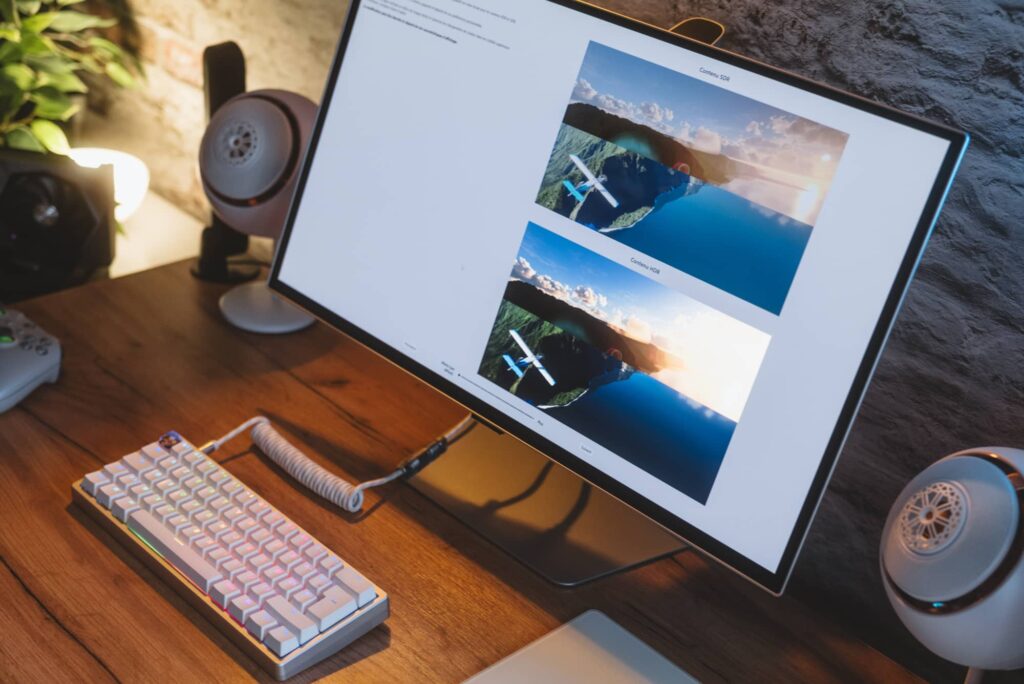 Desktop monitor displaying two airplane photos over coastal landscapes on a wooden desk, with a white mechanical keyboard and speakers nearby.