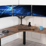 Triple-monitor computer setup on an L-shaped wooden desk with keyboard, mouse, smartphone on a stand, headphones, and a blue mug in a bright office.