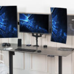 Triple-monitor desk setup on a standing desk: two vertical displays flank a central screen with keyboard, mouse, phone, and laptop nearby.