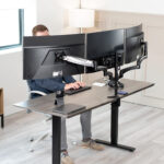 Person seated at a height-adjustable desk with three curved computer monitors mounted on an articulated arm, typing on a keyboard.