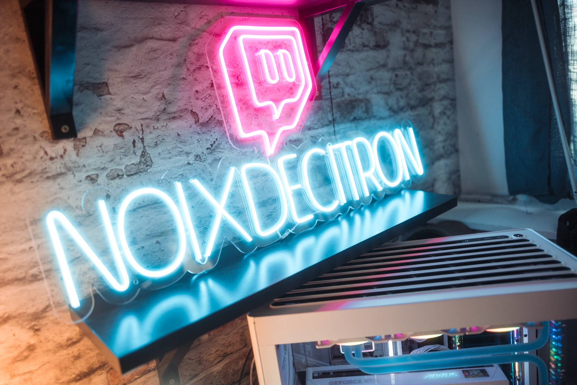 Pink neon sign in a speech-bubble outline above the word 'NOIXDECITRON' on a brick wall, with cool blue neon glow and a computer tower beneath.