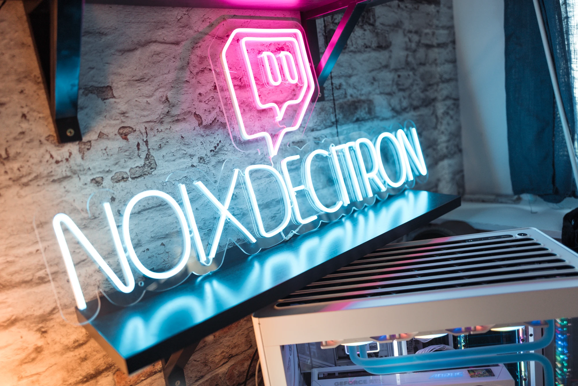 Pink neon speech-bubble logo with the text NOIXDECITRON in blue neon on a brick wall, with a server rack beneath.
