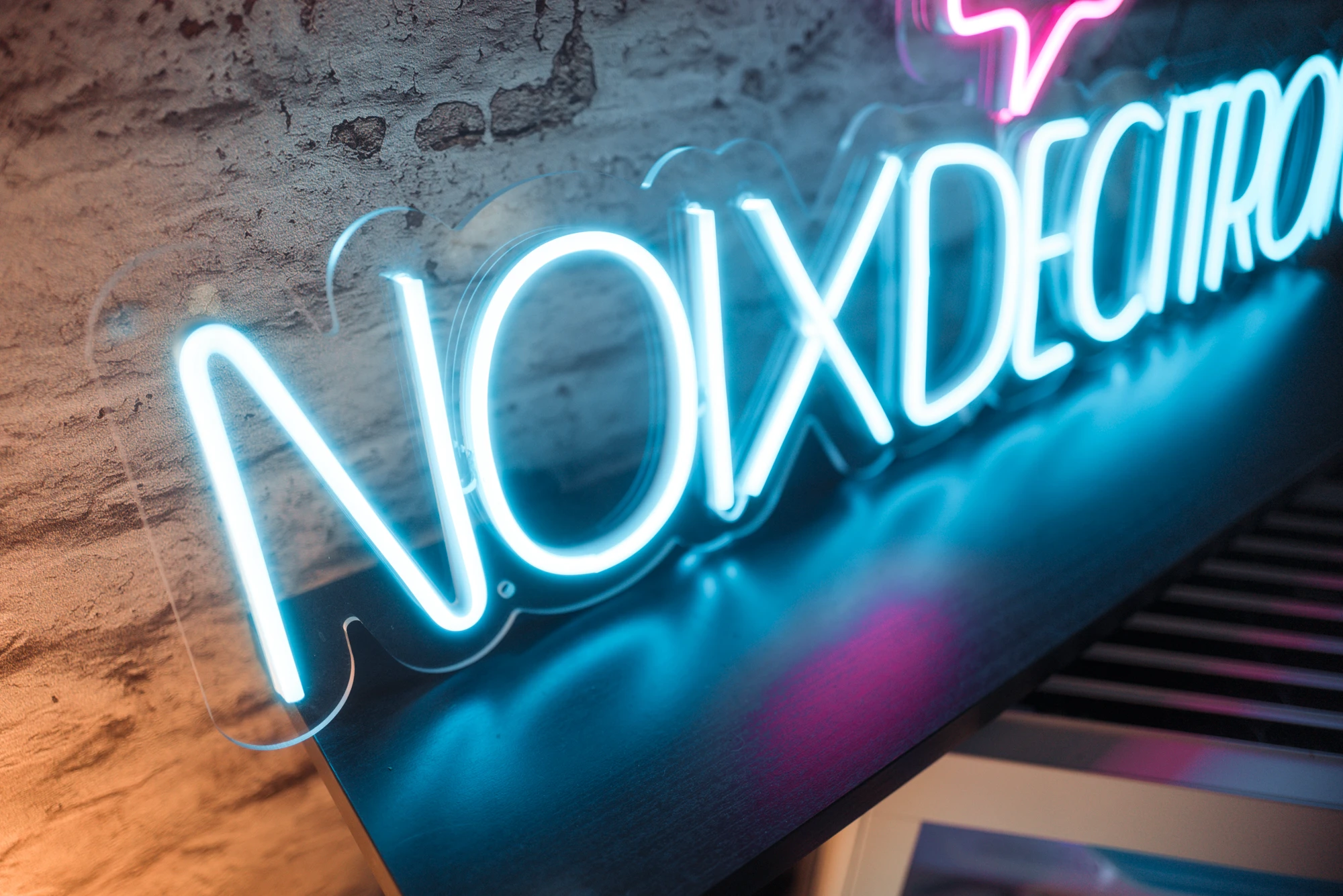 Blue neon sign on a rough brick wall, neon glow reflecting on a piano below (partial word visible).