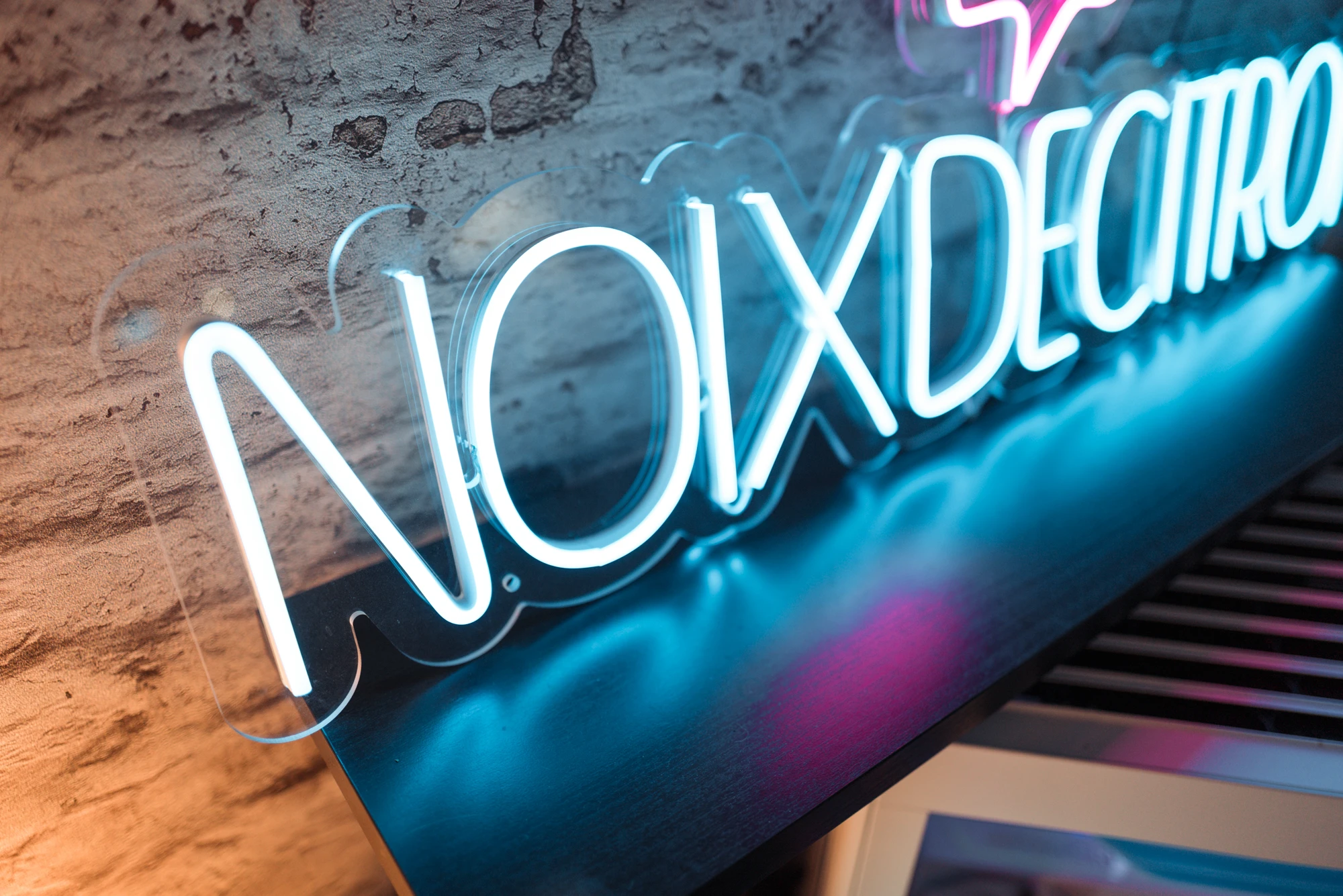 Blue and pink neon sign mounted on a rough brick wall, viewed at a diagonal angle with glowing tubing catching reflections on a dark surface behind it.