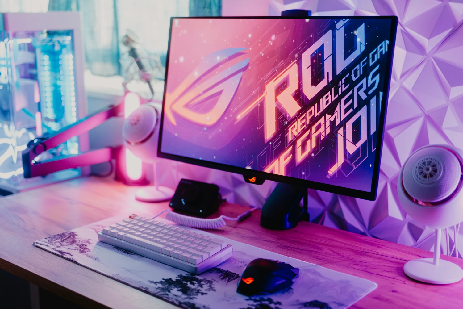 Curved monitor displaying 'ROG Republic of Gamers' with pink-purple neon lighting, keyboard and mouse on a wood desk.