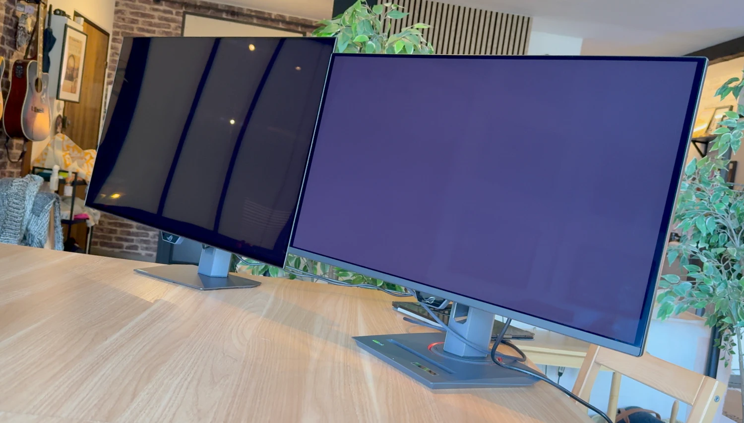 Two wide computer monitors on a shared desk mount in a bright home office; visible cables underneath, with a brick wall, guitars on a rack, and leafy plants in the background.